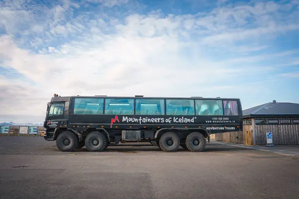 stock image A specialized six-wheeled adventure tour bus by Mountaineers of Iceland parked on a paved surface, with a clear blue sky and a small building in the background.