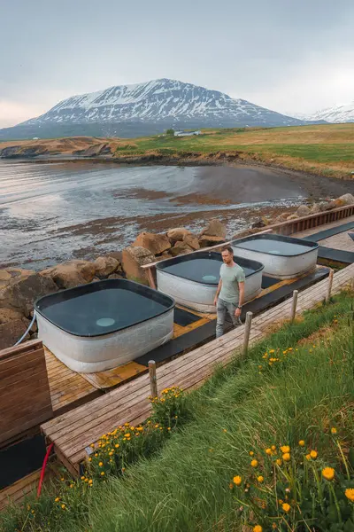 stock image A tranquil Icelandic spa featuring three hot tubs on wooden platforms, surrounded by rocky shores, grassy fields, and yellow wildflowers with a majestic mountain backdrop.