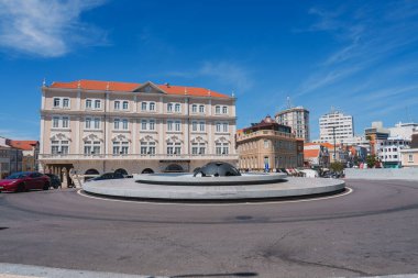 Portekiz 'in Aveiro şehrinde bulunan pitoresk bir kavşakta modern bir çeşme, kırmızı kiremitli çatısı ve açık mavi gökyüzü olan bej renkli çok katlı bir bina yer alıyor..