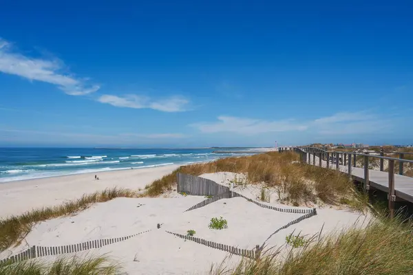 stock image Expansive sandy shore and clear blue waters in Aveiro, Portugal. Elevated wooden boardwalk runs parallel to the beach, with sand dunes and gentle ocean waves.