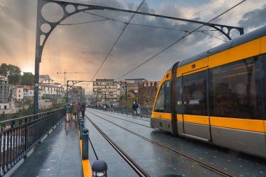 Porto, Portekiz 'deki Dom Luis Köprüsü' nden dramatik bir günbatımında bir tramvay geçer. Islak yüzey ve tarihi binalar yansıtıcı, atmosferik bir manzara yaratıyor..