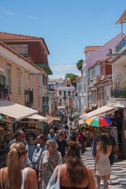 Portekiz, Cascais 'de canlı bir cadde, insanların alışveriş yaptığı, balkonlu renkli binalar ve berrak mavi gökyüzünün altında parke şeklinde kaldırımlar..