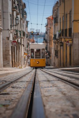 Çekici sarı bir tramvay Portekiz, Lizbon 'da dar, kaldırım taşı bir caddede yol alıyor. Balkonları ve sokak lambaları olan renkli binalar, arka planda Tagus Nehri olan yol boyunca uzanır..