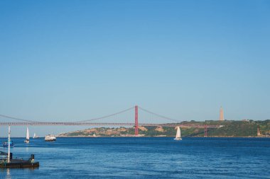 25 de Abril Köprüsü, Portekiz 'in Lizbon kentindeki Tagus Nehri boyunca uzanır ve arka planda Kral heykeli İsa' nın Sığınağı bulunur. Yelkenliler ve feribot sakin sularda yol alır..