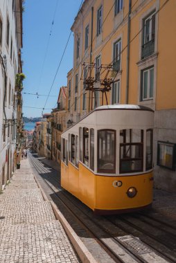 Canlı sarı bir tramvay Lizbon, Portekiz 'de dar, parke taşlı bir caddede yol alır. Pastel renkli binalar ve arka planda Tagus Nehri' ni görür..