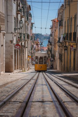 Klasik bir sarı tramvay Lizbon, Portekiz 'de dik ve dar bir caddede yol alır. Demirden yapılma balkonları olan geleneksel Avrupa binaları, arka planda Tagus Nehri olan cadde boyunca uzanır..