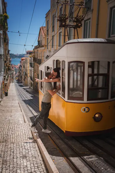 stock image Charming street scene in Lisbon, Portugal, featuring a couple sharing a romantic moment on the iconic yellow Elevador da Bica tram, with a glimpse of the Tagus River.