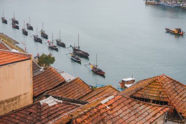 Portekiz 'in Porto kentindeki Douro Nehri' nin sakin bir sahnesinde geleneksel Rabelo tekneleri ve bulutlu gökyüzünün altında kendine özgü kırmızı kiremitli çatılar yer alıyor..