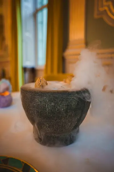 stock image A dark stone bowl emitting misty vapor on a white tablecloth, set in a high-end dining venue with gold accents and tall windows draped with yellow curtains.