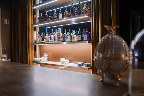 Stock image A sophisticated bar interior featuring a well-organized display of liquor bottles on illuminated shelves, elegant golden curtains, and a decorative Faberge egg.