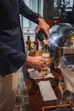 Siyah ceketli ve açık renk pantolonlu biri lüks bir otelde flüt bardağına şampanya döker. Masada altın bir buz kovası, beyaz peçeteler ve ayrıntılara dikkat çeken güneş gözlükleri yer alıyor..