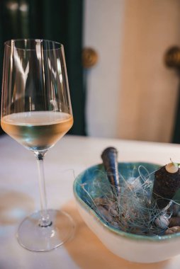 Bir bardak beyaz şarap ve seramik kasede gurme aperatifi bulunan sofistike yemek sahnesi. Lüks dekor ve sanatsal sunum üst düzey ambiyansı vurguluyor.