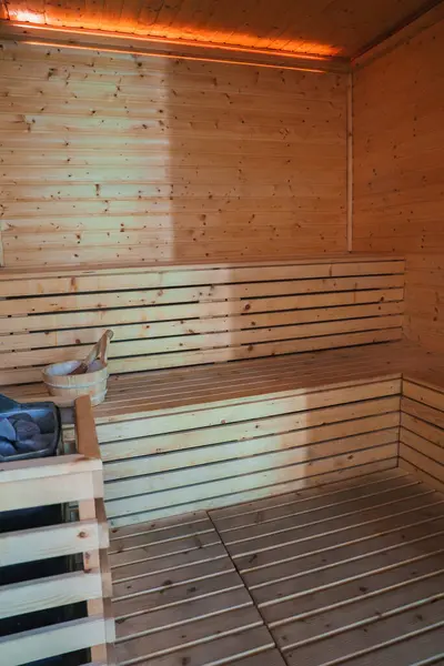 stock image Experience relaxation in a high-end sauna room featuring light wood paneling, benches, and essential steam tools. Warm lighting enhances the cozy atmosphere.