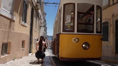 Klasik bir sarı tramvay Lizbon, Portekiz 'de dik ve dar bir caddede yol alır. Geleneksel binalar ve Tagus Nehri 'nin uzak mavi suları manzarayı güzelleştirir..