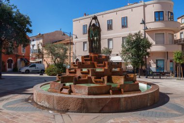 Akdeniz binaları ve açık mavi gökyüzünün altındaki ağaçlarla çevrili, Sardunya meydanında bronz heykeli olan katmanlı taş bir çeşme..