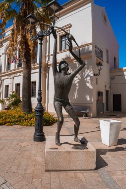 İtalya 'nın Sardinya kentindeki güneşli bir plazada bronz bir heykel yukarı doğru uzanır. Sahnede klasik bir sokak lambası, beyaz bir Akdeniz binası ve bir palmiye ağacı yer alıyor..