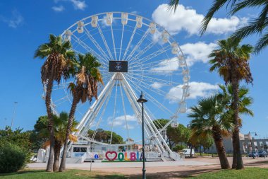 Olbia, Sardinya, İtalya 'da berrak mavi bir gökyüzüne kurulmuş canlı bir dönme dolap. Etrafı bereketli palmiye ağaçları ile çevrili. Modern eğlenceyi doğal güzellikle harmanlıyor..