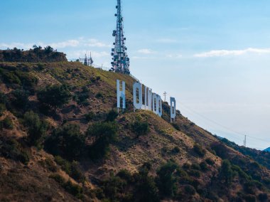 Hollywood tabelası, arkasında iletişim kulesi olan bir yamaç tarafından kısmen gizlenmiş. Kuru arazi ve açık mavi gökyüzü manzarayı belirler.