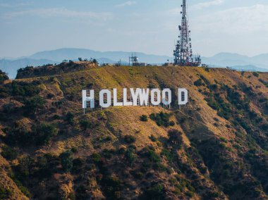 Hollywood tabelası engebeli tepelerde bir iletişim kulesiyle sergileniyor. Uzak dağlar ve berrak mavi bir gökyüzü manzaralı bir zemin oluşturuyor..