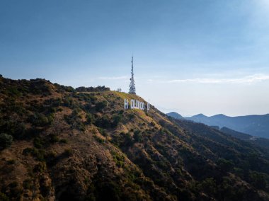 İkonik Hollywood tabelası Los Angeles 'ın engebeli bir yamacında arkasında bir radyo kulesi bulunuyor. Nadir bitki örtüsü ve uzak dağlar görülebilir..