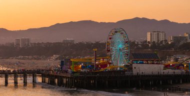 Los Angeles 'taki Santa Monica İskelesi gün batımında insanlarla dolup taşıyor. Dönme dolap, Pasifik Okyanusu 'na ve uzak dağlara karşı parlak bir şekilde aydınlatılır..