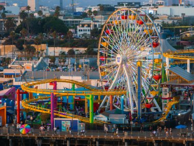 Görüntü Santa Monica Piers Pacific Parkı 'nı çekiyor, dönme dolabı ve hız trenini vurguluyor. Ziyaretçiler şehir manzarasının arkasındaki cazibeyi keşfediyorlar.