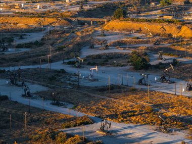 Kaliforniya 'daki bir çöl petrol sahasının geniş bir manzarası. Beton bloknotlar, toprak yollar ve sıcak, altın ışık altındaki elektrik hatları üzerinde sayısız sondaj kulesi var..