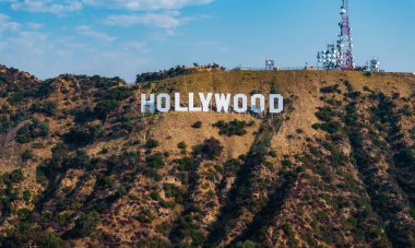 Hollywood tabelası engebeli tepelerde sergileniyor. Arka planda yeşil yamalar, iletişim kuleleri ve Los Angeles 'ta açık mavi bir gökyüzü var..