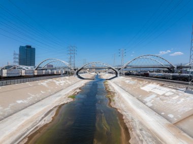 Los Angeles Nehri beton bir kanaldan geçer, kemerli köprülerle geçer. Elektrik hatları ve açık mavi gökyüzüne karşı yükselen yüksek bir bina..