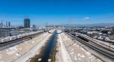 Los Angeles Nehri, altıncı caddedeki viyadük tarafından çerçevelenmiş beton bir kanaldan akar. Los Angeles şehir merkezinde gökyüzü ve yük trenleri görülebilir..