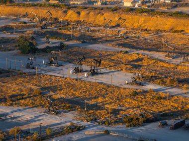 An aerial view captures numerous oil rigs in a California desert. The arid landscape features scrub vegetation and dirt roads, with long shadows cast by the setting sun. clipart