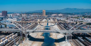 Los Angeles Nehri beton kanalı, geniş kemerli bir köprü, tren rayları, kentsel binalar ve San Gabriel Dağları 'nın hava perspektifi..