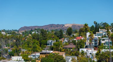 Görüntü, Los Angeles 'taki berrak mavi gökyüzünün altında yeşil ve modern mimari evlerle çevrili tepelerdeki Hollywood tabelasını yakalıyor..