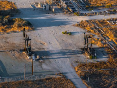 Bir hava görüntüsü Kaliforniya çölünde iki petrol kulesi gösteriyor. Kuru, kumlu arazide seyrek bitki örtüsüyle çevrili boru hatları ve endüstriyel yapılar..