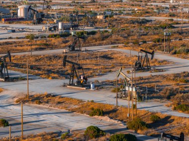 Numerous oil rigs operate in a desert landscape in California, with dusty roads and sparse vegetation. The setting sun casts long shadows over the scene. clipart