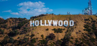 Hollywood tabelası yeşil bantlı engebeli tepelerde sergileniyor. Açık mavi gökyüzünün altında iletişim kuleleri görülebiliyor..
