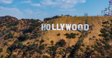Hollywood tabelası Los Angeles, California 'nın engebeli tepelerinde sergilenmektedir. Yakınlarda iletişim kuleleri ve bulutsuz açık bir gökyüzü vardır..