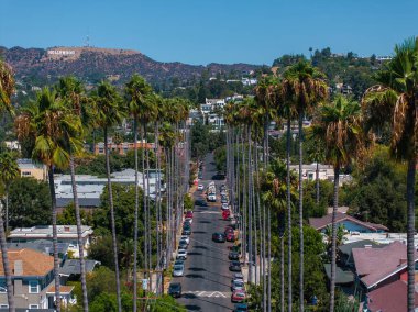 Hollywood, Los Angeles 'ta bir caddede yüksek palmiye ağaçları ve konut binaları var. Tepelerde görünen ikonik Hollywood tabelası açık bir gökyüzünün altında..
