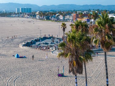 Los Angeles 'taki Venedik Plajı' nda arka planda Santa Monica Dağları 'nın bulunduğu kalabalık bir kaykay parkı, ikonik palmiye ağaçları ve renkli binalar yer alıyor..