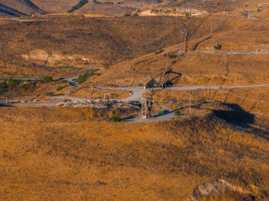 Kaliforniya 'da kuru bir çöl arazisinde petrol kuleleri ve seyrek bitki örtüsü bulunur. Sıcak güneş ışığı engebeli tepeleri ve endüstriyel ekipmanları aydınlatıyor.