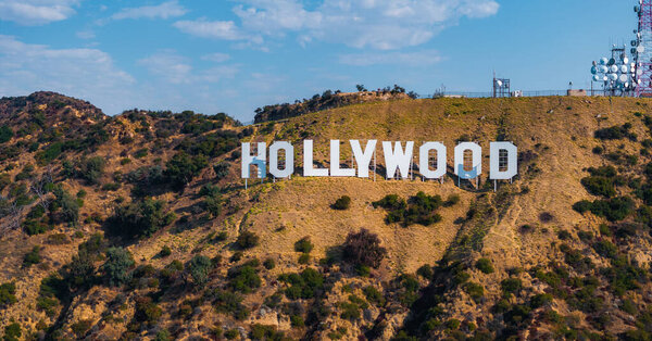 Голливудский знак отображается на холмах Лос-Анджелеса, Калифорния, с башнями связи поблизости и ясным небом с несколькими облаками.