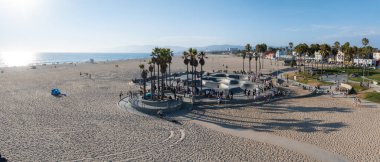 Los Angeles 'taki Venice Beach' te palmiye ağaçları ve ziyaretçilerle çevrili hareketli bir kaykay parkı bulunmaktadır. Kumlu sahil okyanusa kadar uzanır, uzak dağlar görülebilir..