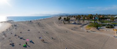 Los Angeles 'taki Venedik Sahili' nde kumlu sahiller, renkli şemsiyeler ve metal bir heykel bulunur. Hurma ağaçları ve dağlar berrak bir gökyüzünün altında görünür..