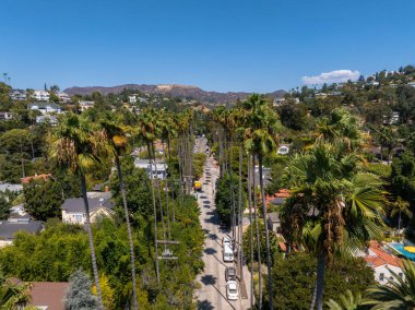 Hollywood, Los Angeles 'ta yüksek palmiye ağaçlarıyla kaplı bir caddenin havadan görünüşü. Sembolik Hollywood tabelası, yeşilliklerle çevrili güneşli tepelerde görülebilir..