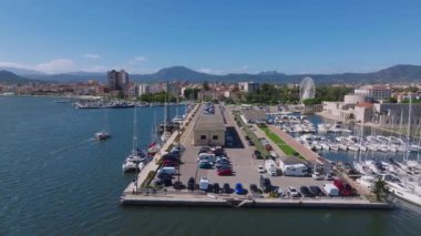 Modern binalar ve berrak mavi suyla çevrili Olbias marinasına bir yelkenli yanaşıyor ve doğal güzelliğin ve şehir hayatının karışımını sergiliyor..