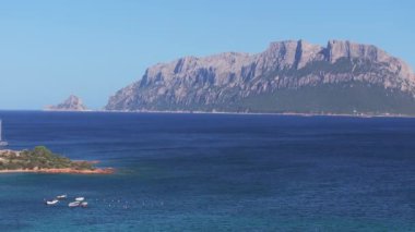 Sardinya 'nın turkuaz suları, küçük tekneleri, yemyeşil alanları, dağınık evleri ve arka planda yuvarlanan tepeleri olan havadan manzaralı bir koy..
