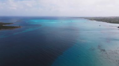 Sardunya sahilinin turkuvaz suları, kumlu kıyı şeridi ve plaja gidenlerle hava manzarası. Kıyı şeridinde bir yol, park edilmiş arabalar ve yemyeşil alanlar yer alıyor..