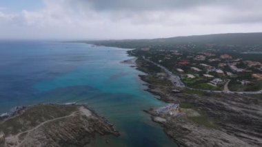 Sardunya sahilinin turkuvaz suları, kumlu kıyı şeridi ve plaja gidenlerle hava manzarası. Kıyı şeridinde bir yol, park edilmiş arabalar ve yemyeşil alanlar yer alıyor..
