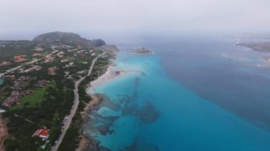 Sardunya sahilinin turkuvaz suları, kumlu kıyı şeridi ve plaja gidenlerle hava manzarası. Kıyı şeridinde bir yol, park edilmiş arabalar ve yemyeşil alanlar yer alıyor..