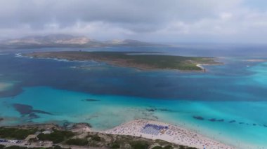 Sardunya sahilinin turkuvaz suları, kumlu kıyı şeridi ve plaja gidenlerle hava manzarası. Kıyı şeridinde bir yol, park edilmiş arabalar ve yemyeşil alanlar yer alıyor..